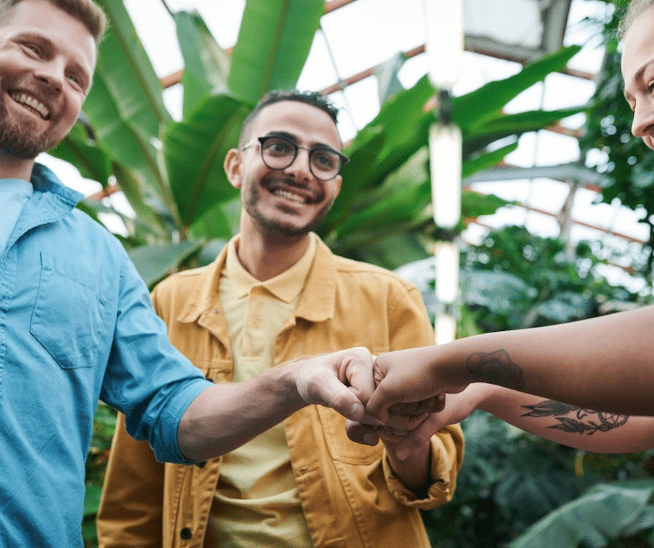 Three supportive friends fist tap in a greenhouse, smiling and radiating a 'we're here for you' vibe.