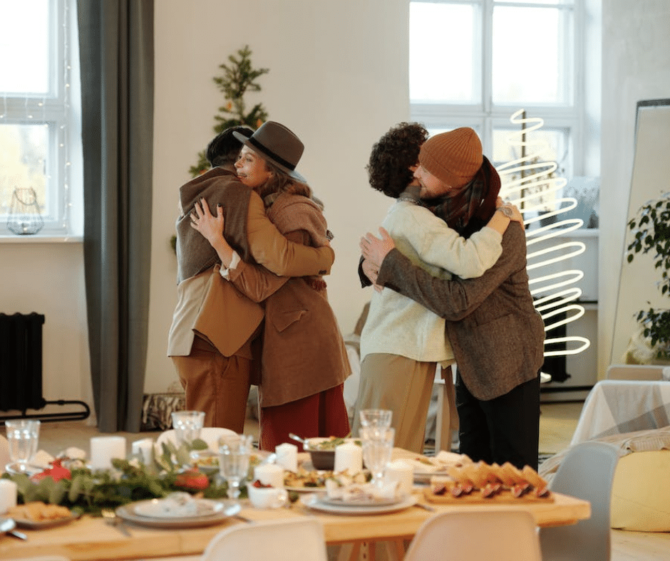 A supportive group of four individuals share heartfelt hugs next to a dinner table, embodying gratitude and unity in their recovery journey.