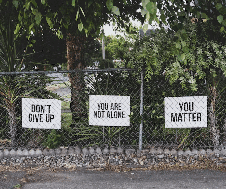 Side view of a lawn with trees and plants lining a fence. Three lawn signs display motivational quotes: 'Don't Give Up,' 'You Are Not Alone,' and 'You Matter.'