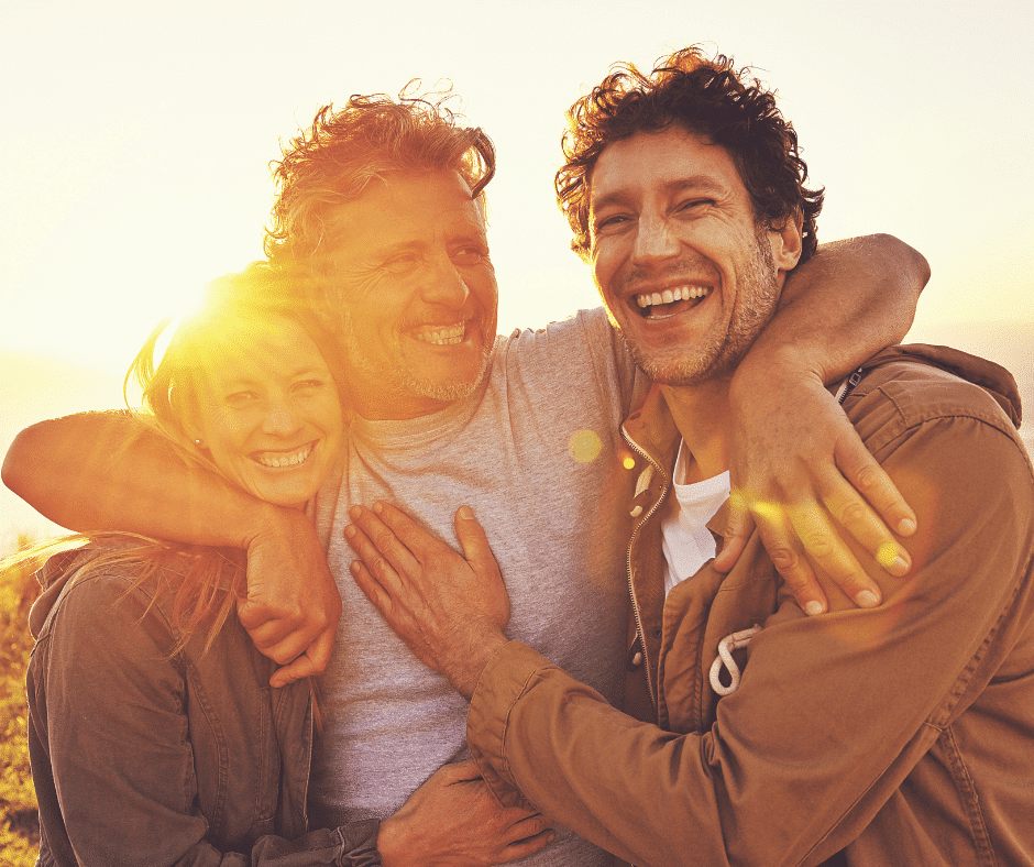 Three friends embracing during Recovery Month with arms around each other, smiling, with the sun gleaming brightly behind them.