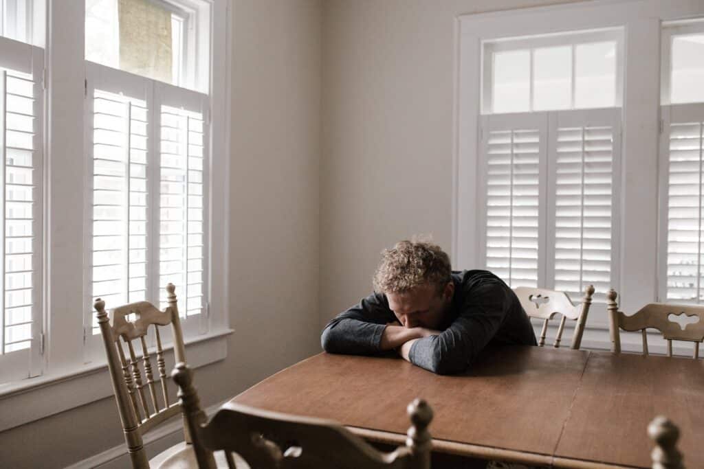 A man appearing distressed and deep in thought, with his head resting on his hands over a dining room table.