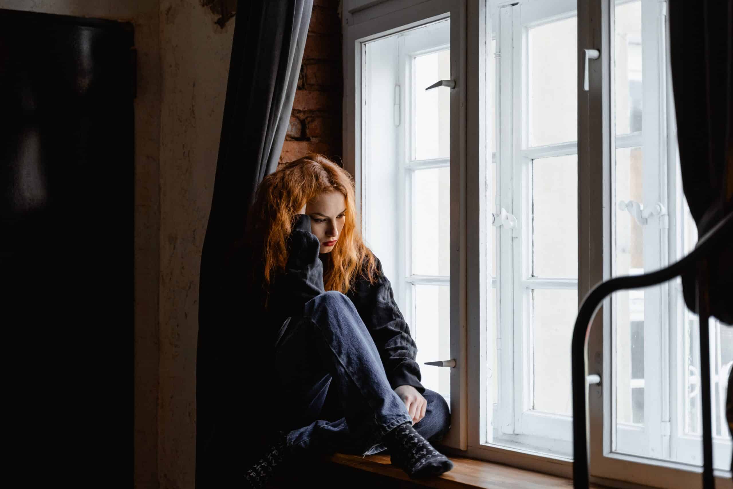 Woman sitting on a window sill, deep in thought with a sad expression, reflecting on the challenges of opioid use disorder.