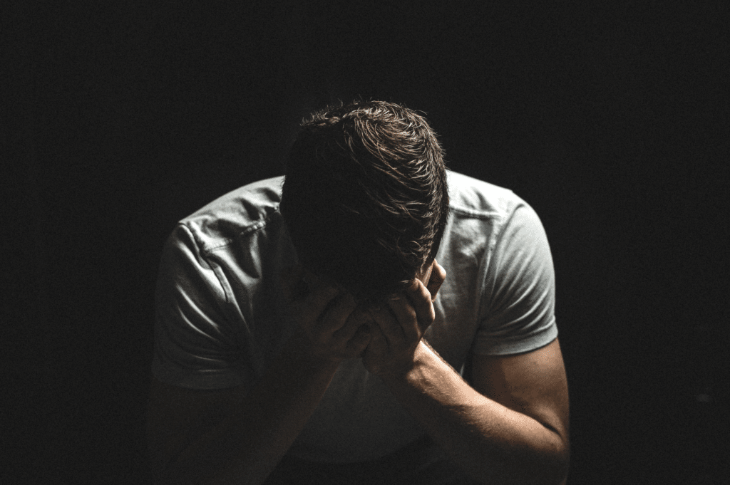 A man in a dark room with his head in his hands, conveying the sense of distress and low self-efficacy.